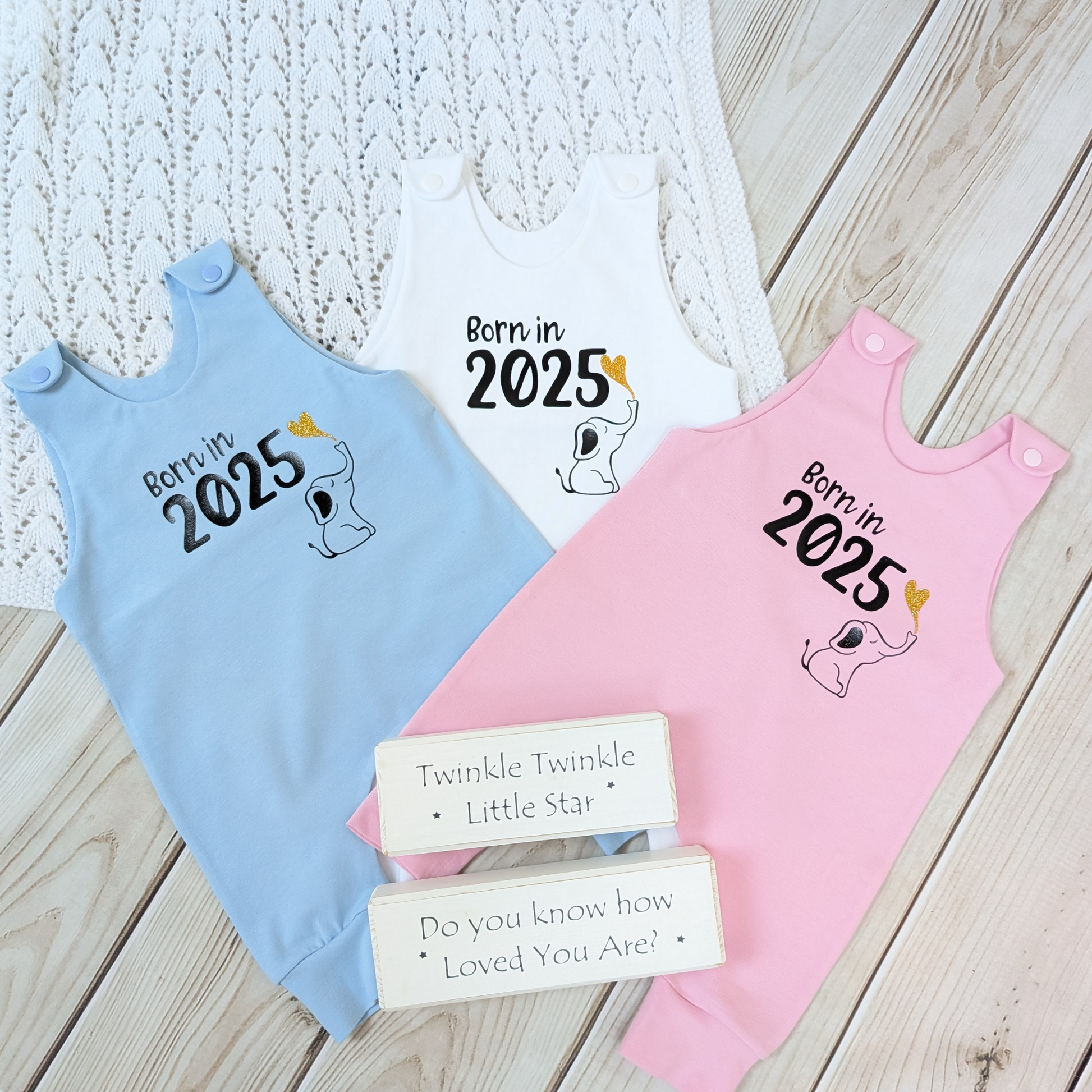 Flatlay-three-rompers-in-pink-white-and-blue-long-leg-romper-with-phrase-born-in-2025-on-front-along-with-cute-elephant-blowing-a-glitter-heart-bubble-blanket-pom-poms-wooden-blocks-for-props