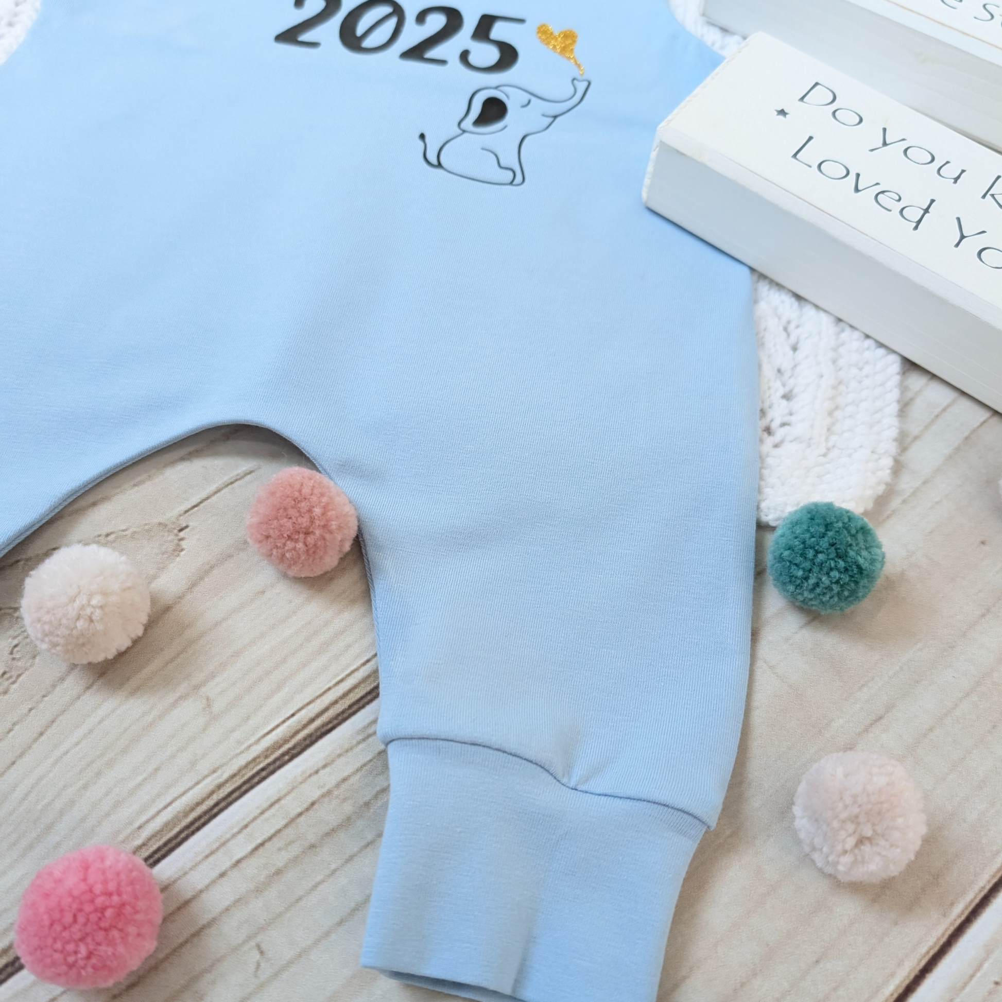 Close-up-leg-Flatlay-blue-long-leg-romper-with-phrase-born-in-2025-on-front-along-with-cute-elephant-blowing-a-glitter-heart-bubble-blanket-pom-poms-wooden-blocks-for-props