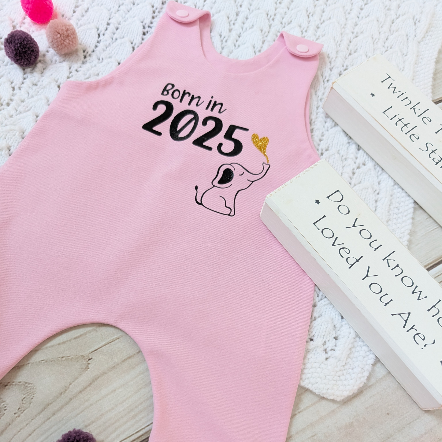 Flatlay-pink-long-leg-romper-with-phrase-born-in-2025-on-front-along-with-cute-elephant-blowing-a-glitter-heart-bubble-blanket-pom-poms-wooden-blocks-for-props