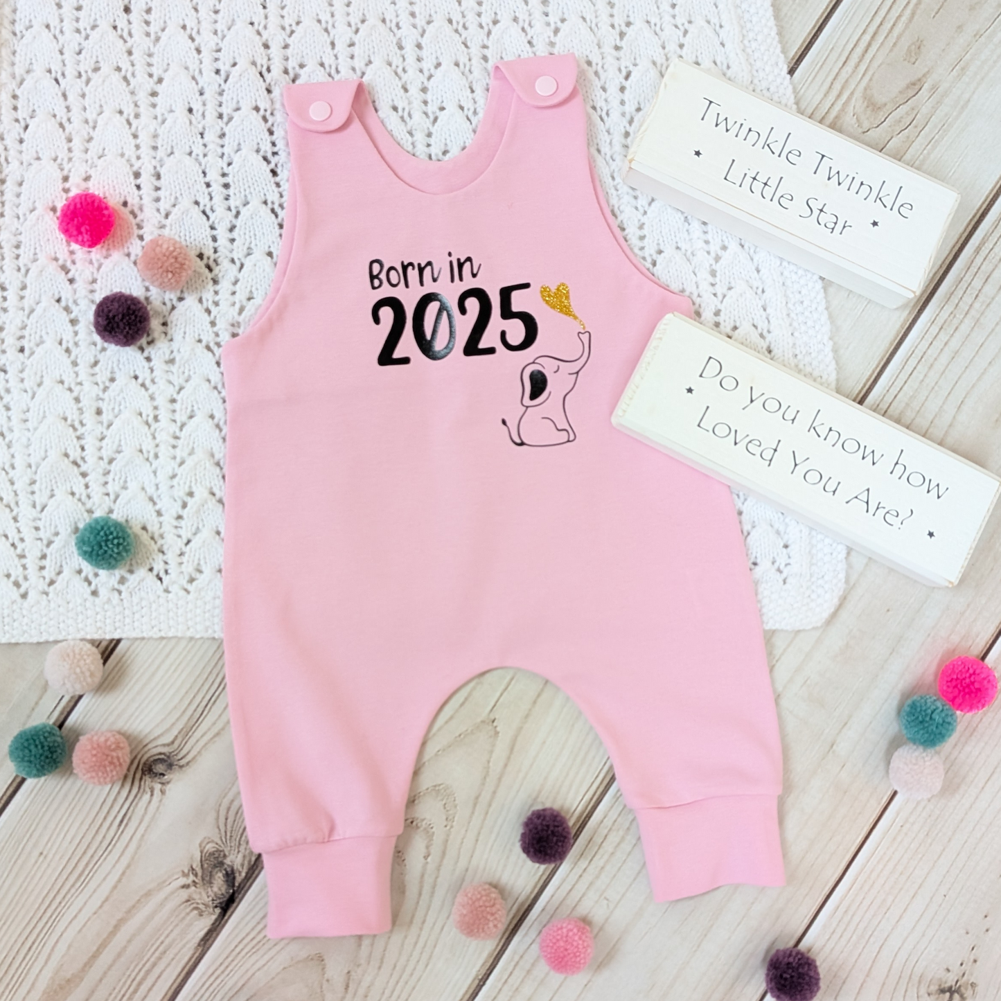 Flatlay-pink-long-leg-romper-with-phrase-born-in-2025-on-front-along-with-cute-elephant-blowing-a-glitter-heart-bubble-blanket-pom-poms-wooden-blocks-for-props