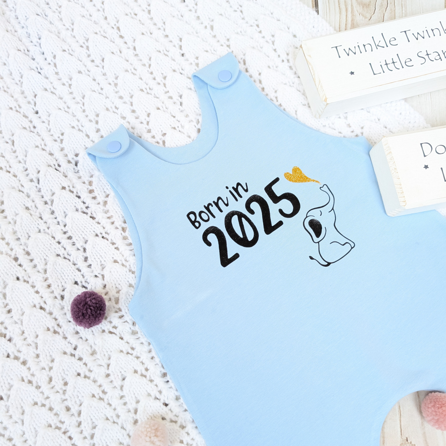 Flatlay-blue-long-leg-romper-with-phrase-born-in-2025-on-front-along-with-cute-elephant-blowing-a-glitter-heart-bubble-blanket-pom-poms-wooden-blocks-for-props