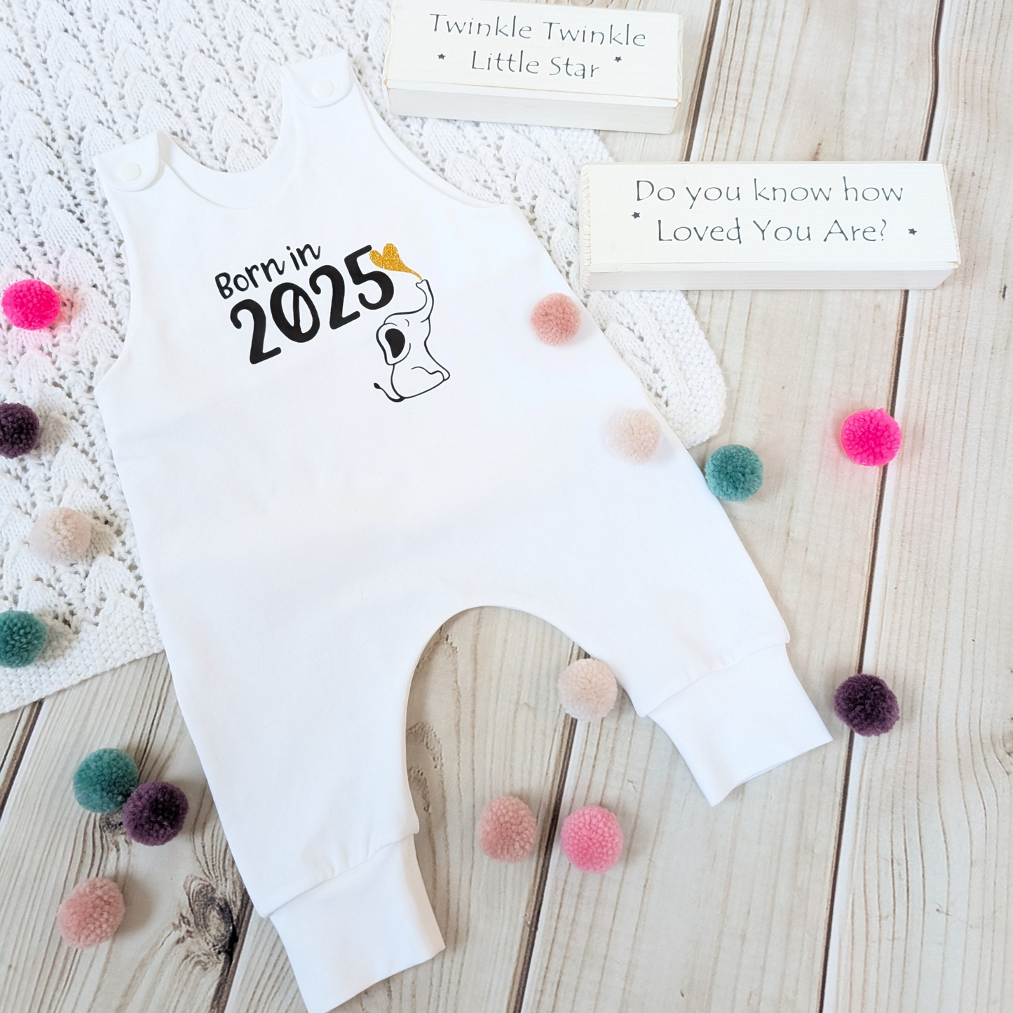 Flatlay-white-long-leg-romper-with-phrase-born-in-2025-on-front-along-with-cute-elephant-blowing-a-glitter-heart-bubble-blanket-pom-poms-wooden-blocks-for-props