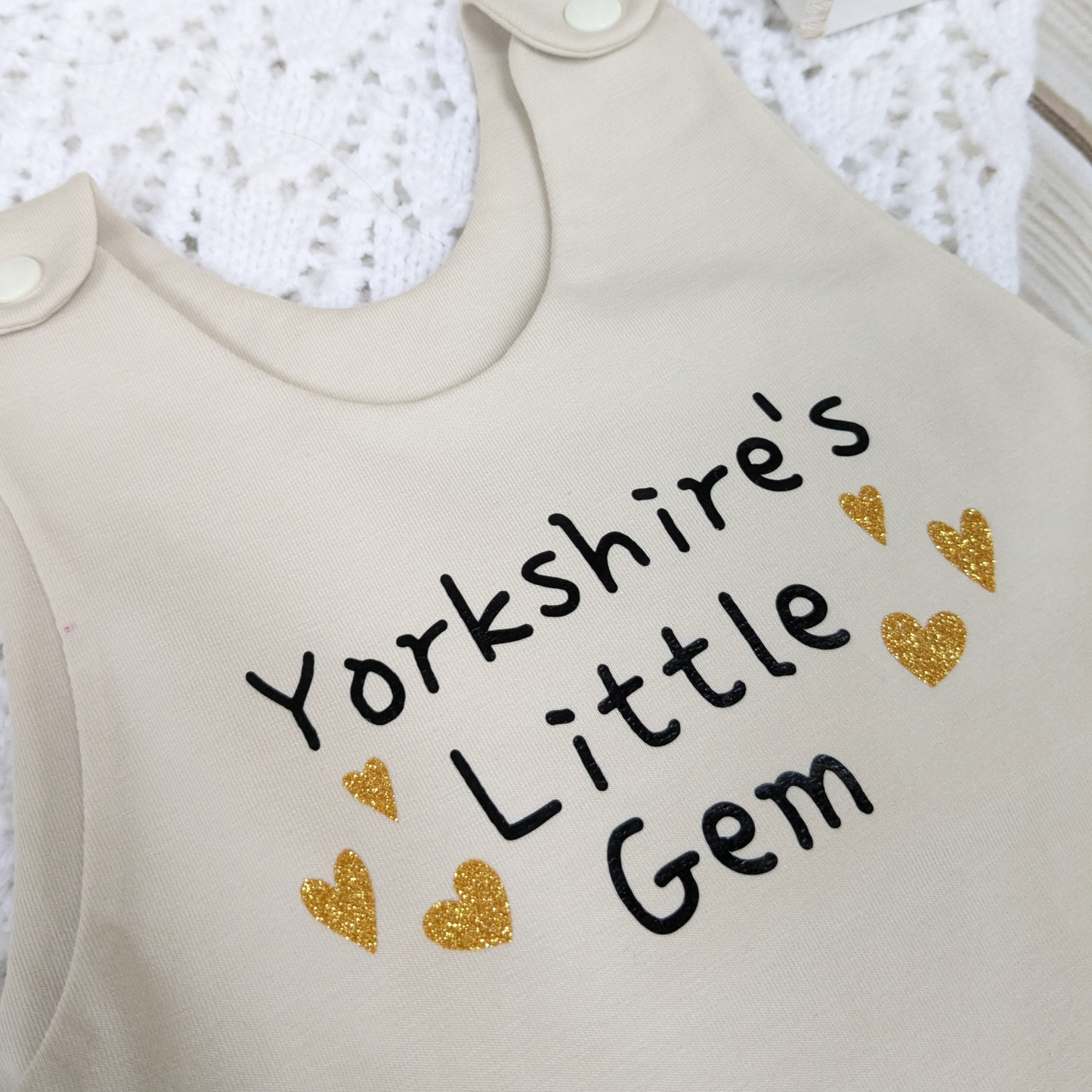 close up-neutral-beige-long-leg-romper-with-phrase-yorkshire's-little-gem-gold-hearts-front-beige-shoulder-snaps-photo-props